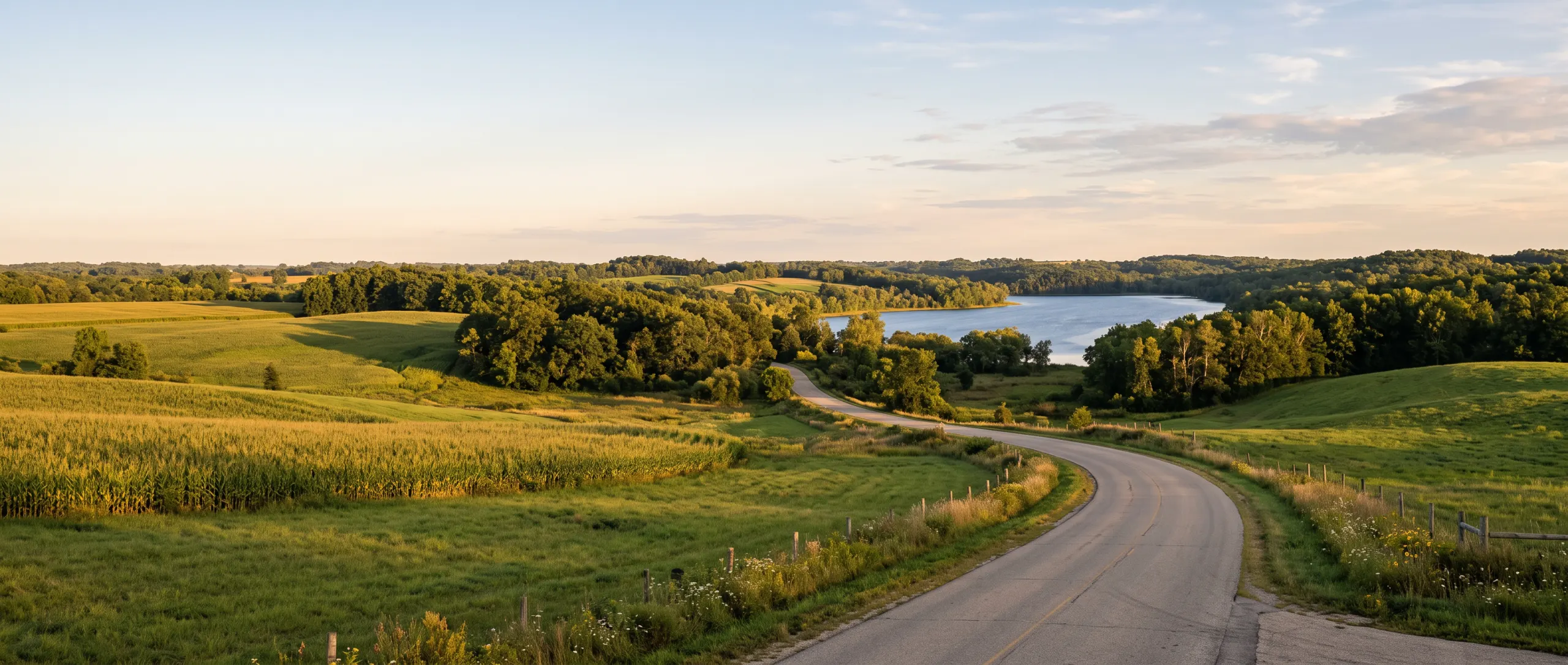 A quiet country road curving past cornfields and trees toward a blue lake at golden hour